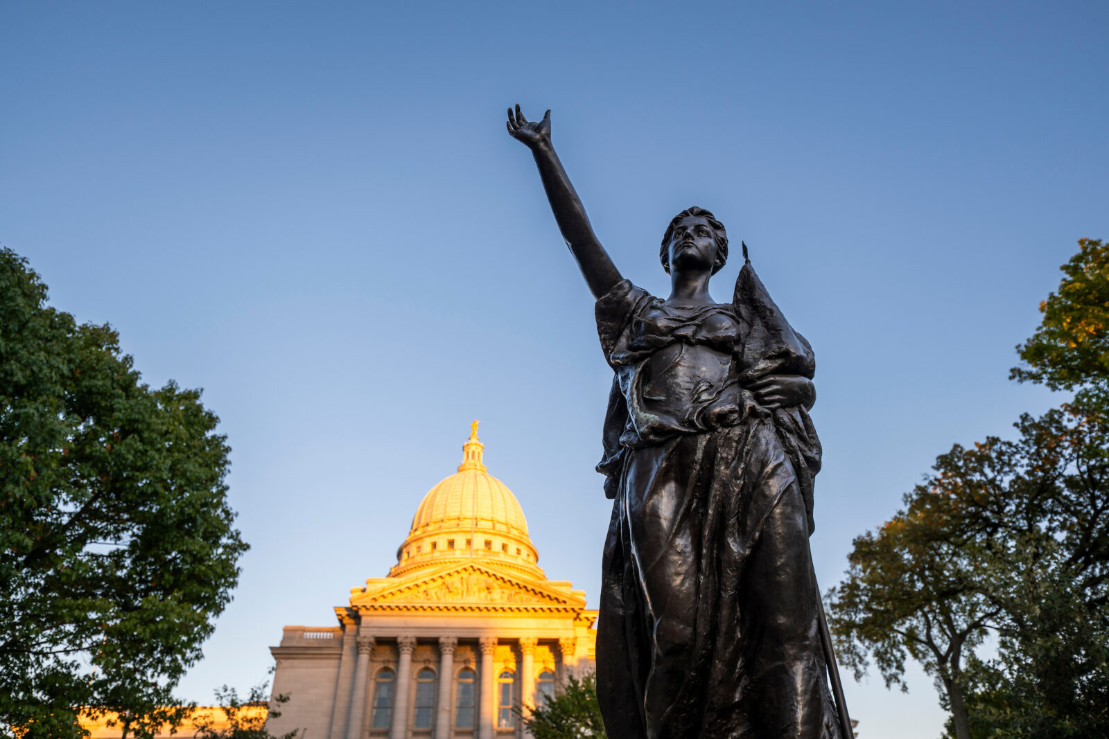 The Forward statute frames a view of sunset light shining on the dome of the Wisconsin State Capitol building near the University of Wisconsin–Madison campus